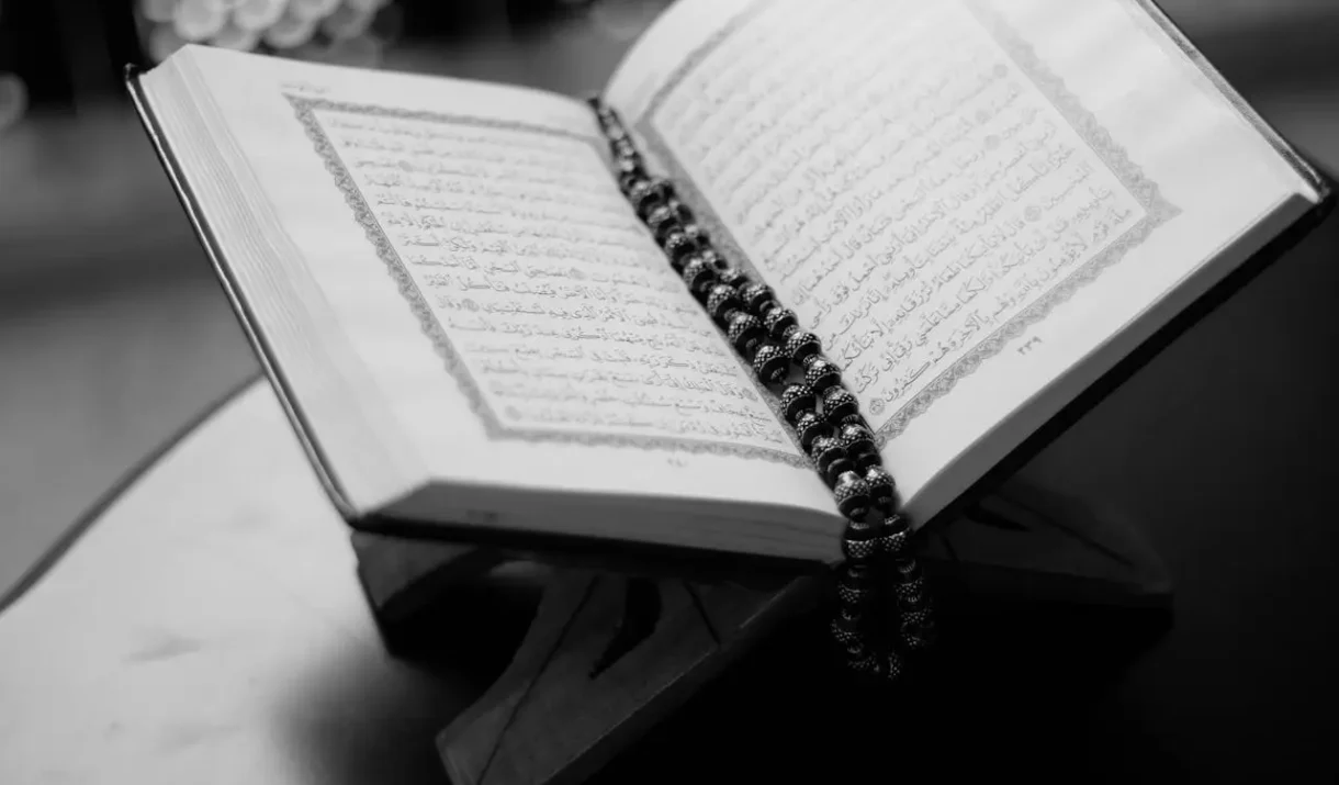 A black and white close-up of an open Quran on a wooden stand with prayer beads, symbolizing Islamic faith and devotion.
