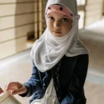 Portrait of a young girl in a hijab reading the Quran indoors. The scene captures a peaceful, cultural moment.