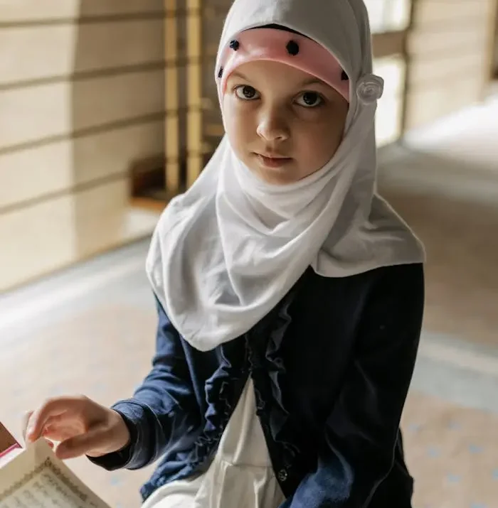 Portrait of a young girl in a hijab reading the Quran indoors. The scene captures a peaceful, cultural moment.